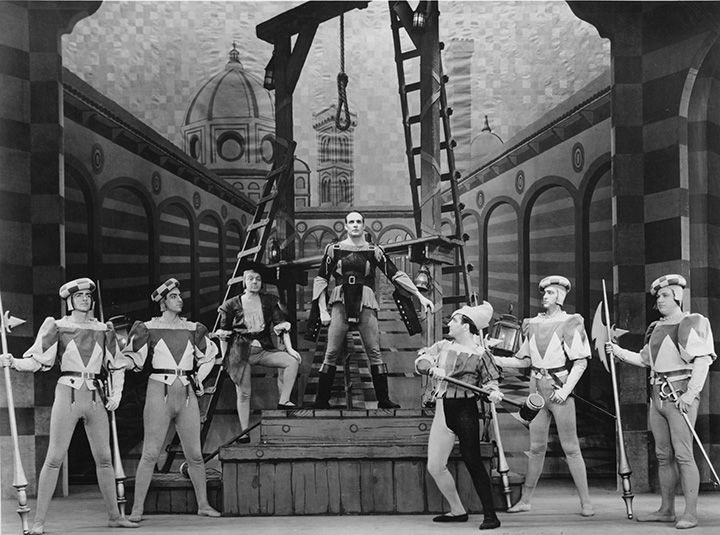 Benvenuto Cellini standing beneath the gallows, surrounded by armed men. Behind him, a stage backdrop of the Duomo and other landmarks of Florence.