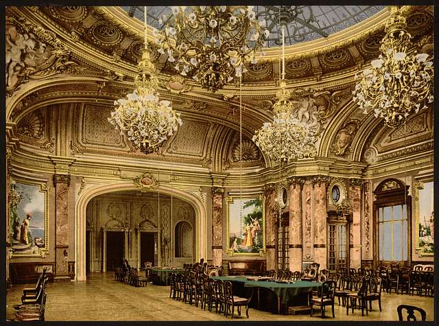 An opulent room, with three chandeliers and a lot of gold. A long gaming table is at center-right.